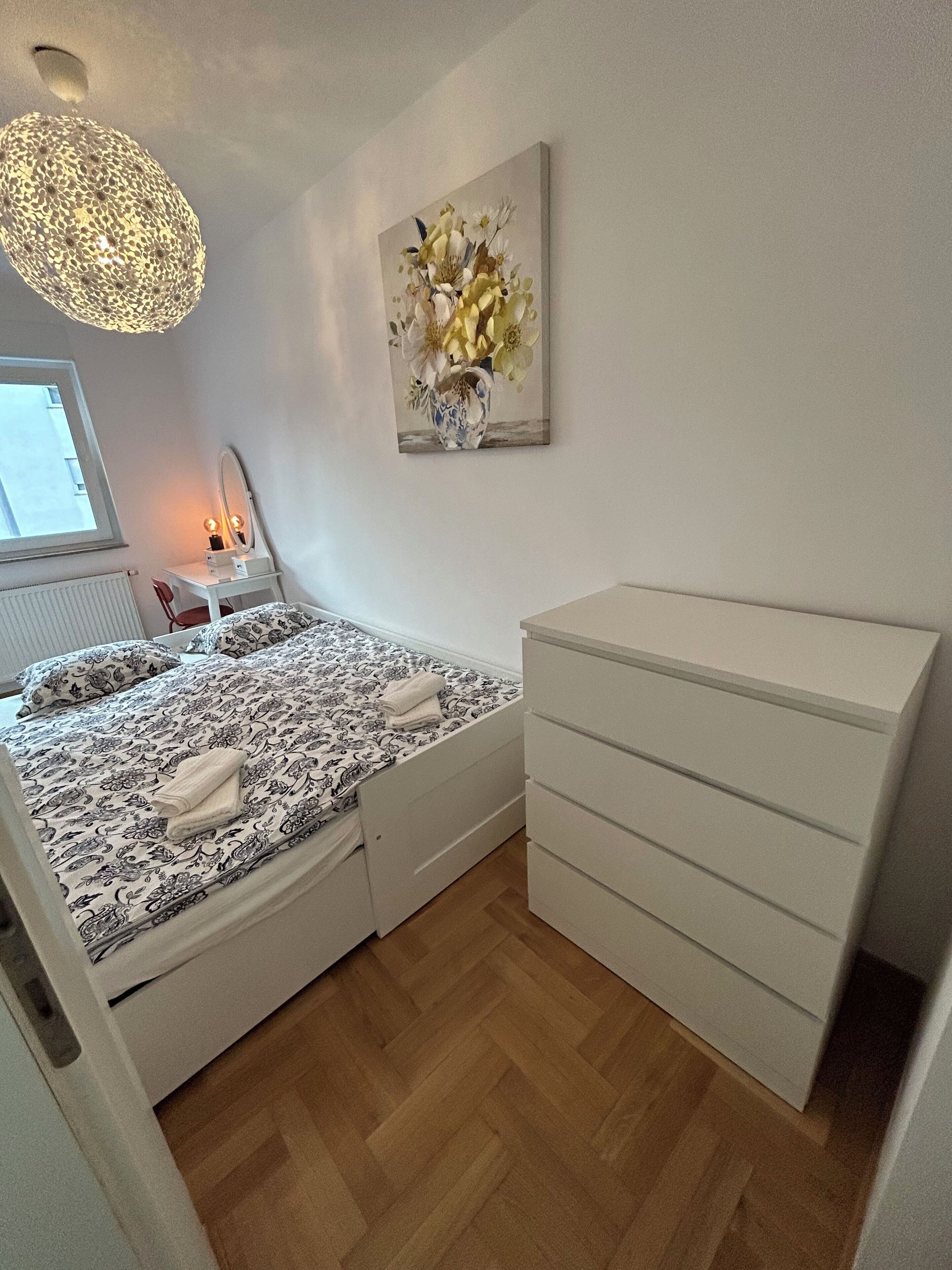 A bedroom with a bed, a dresser, a desk with a mirror, and a wall-mounted artwork.