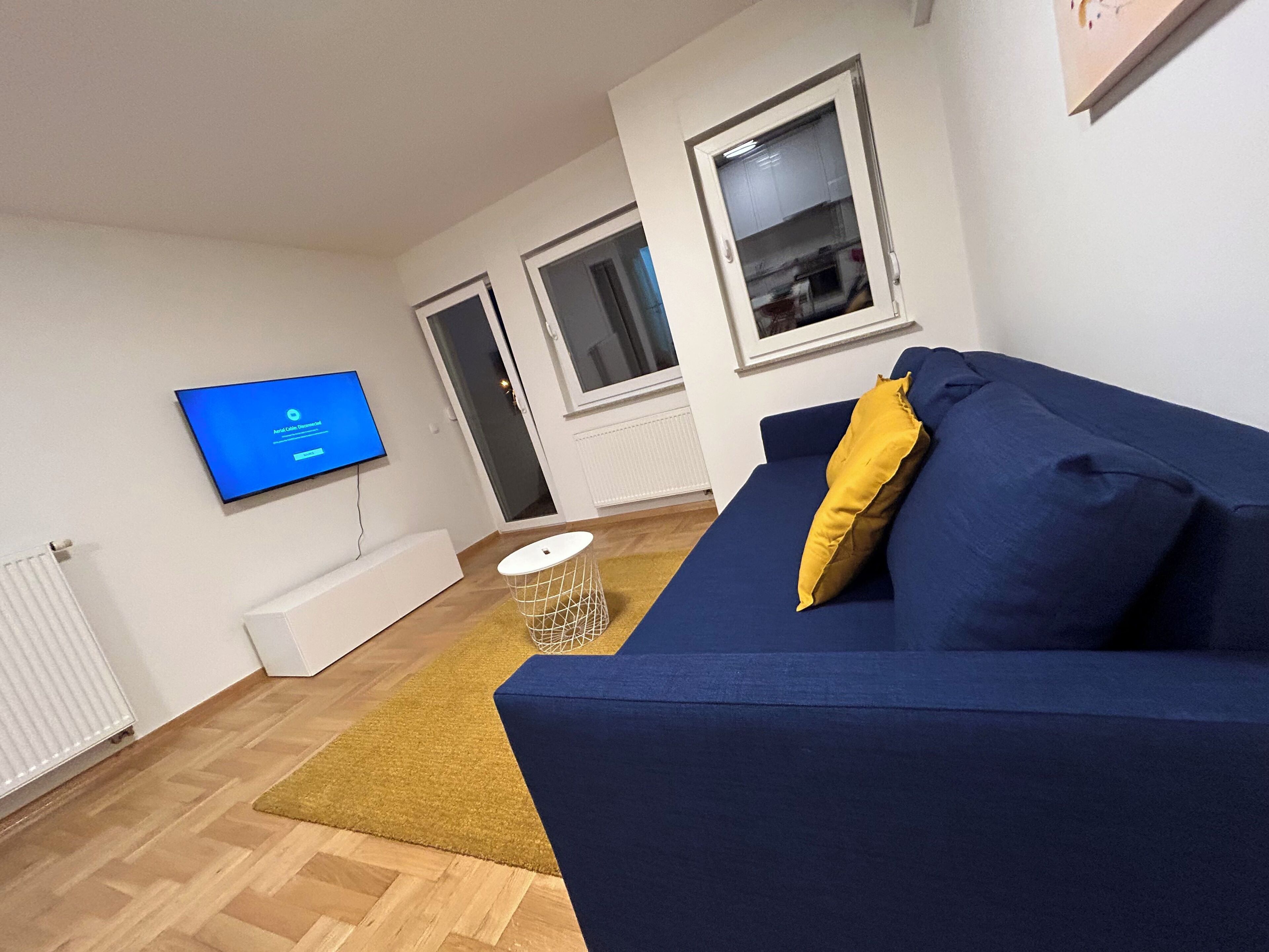 Un salón con un sofá azul, un cojín amarillo, un televisor en la pared y un banco blanco.