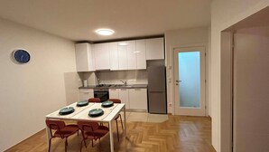 A compact kitchen with white cabinets, a stove, and a refrigerator. There is a dining table with four chairs and a clock on the wall. - Apartment Fran (Zagreb)