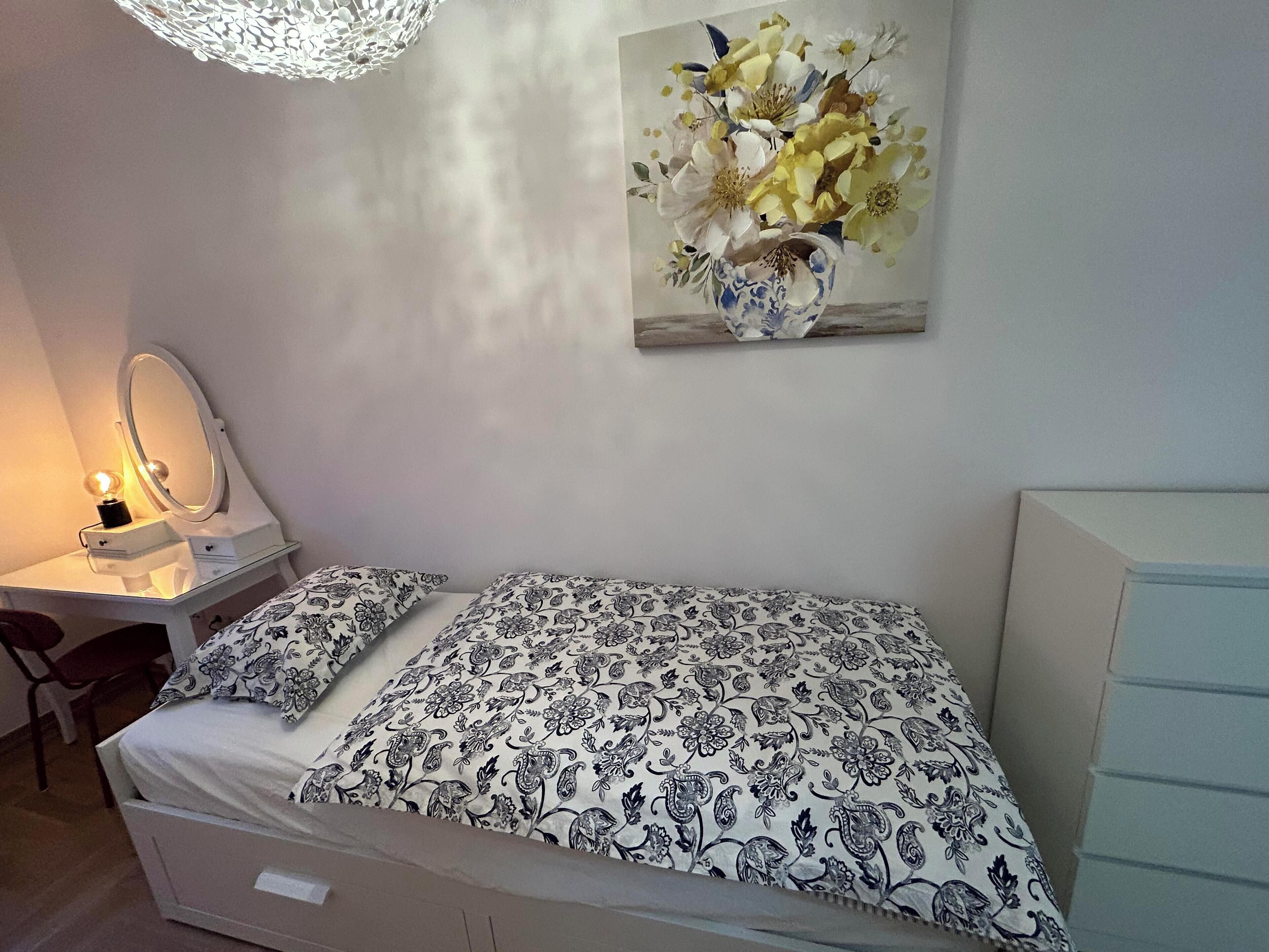 A bedroom with a bed, a bedside table with a lamp, a mirror, and a wall-mounted painting.