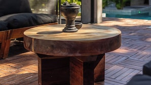 Een moderne buitenruimte met een ronde houten tafel en een zwarte stoel met kussens, met uitzicht op een zwembad en weelderige begroeiing.