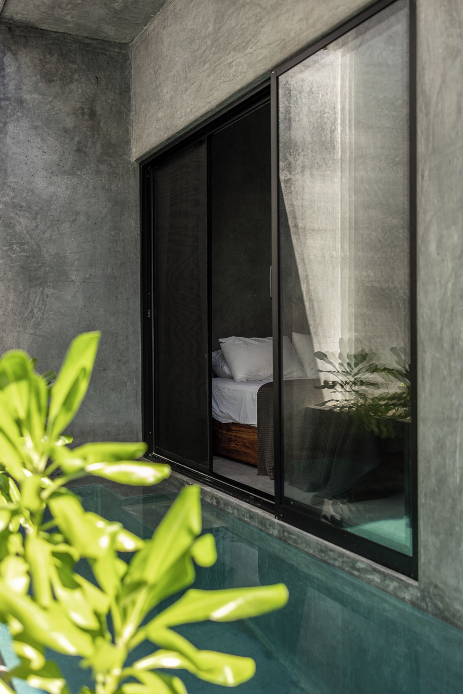 Ein modernes Zimmer mit einer großen SchiebeglasTür, die auf einen Balkon führt, einem Bett mit weißen Bettwäsche und einer kleinen Pflanze auf dem Boden.