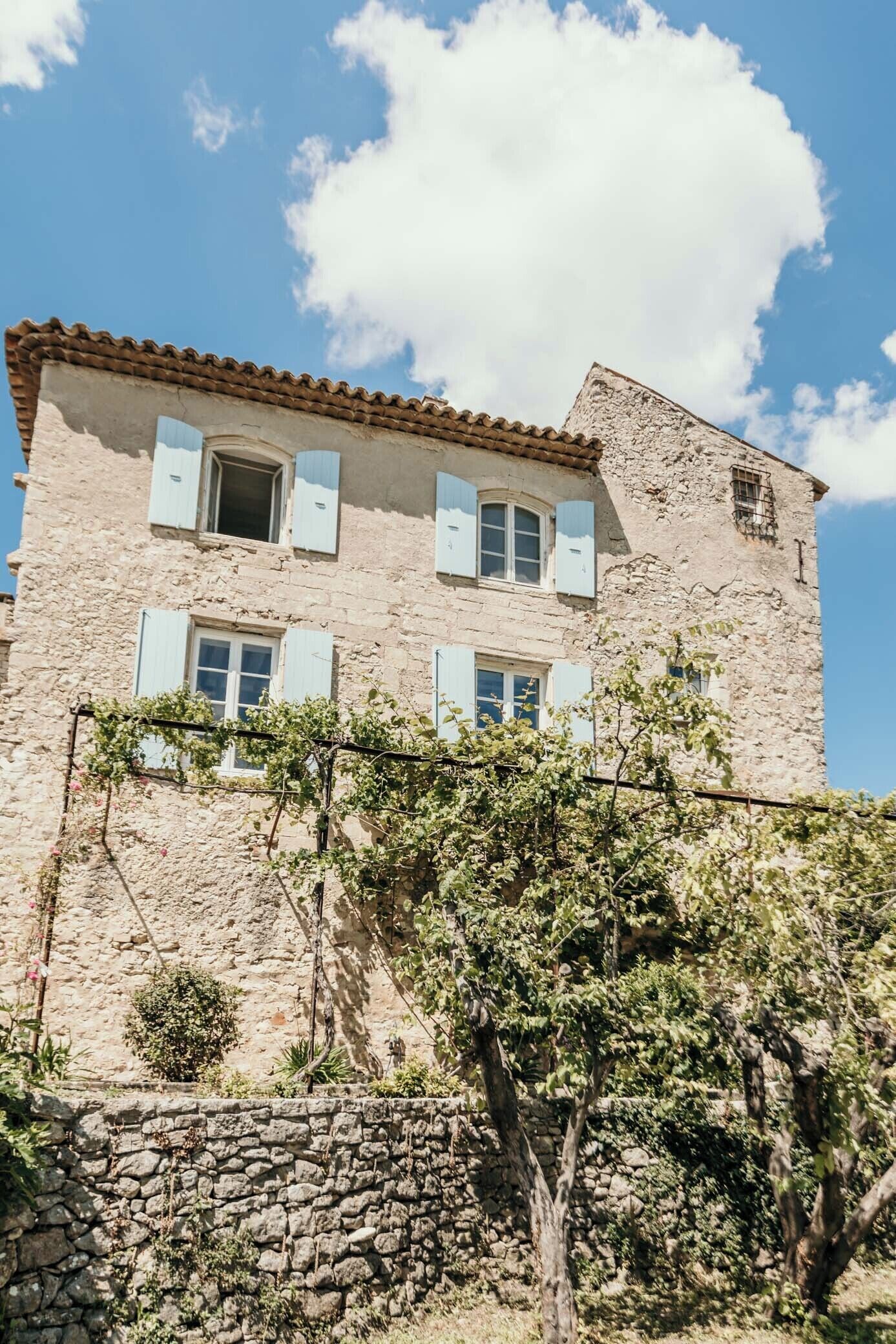Un bâtiment en pierre avec un toit en tuiles, de nombreuses fenêtres avec volets et un mur recouvert de vigne.