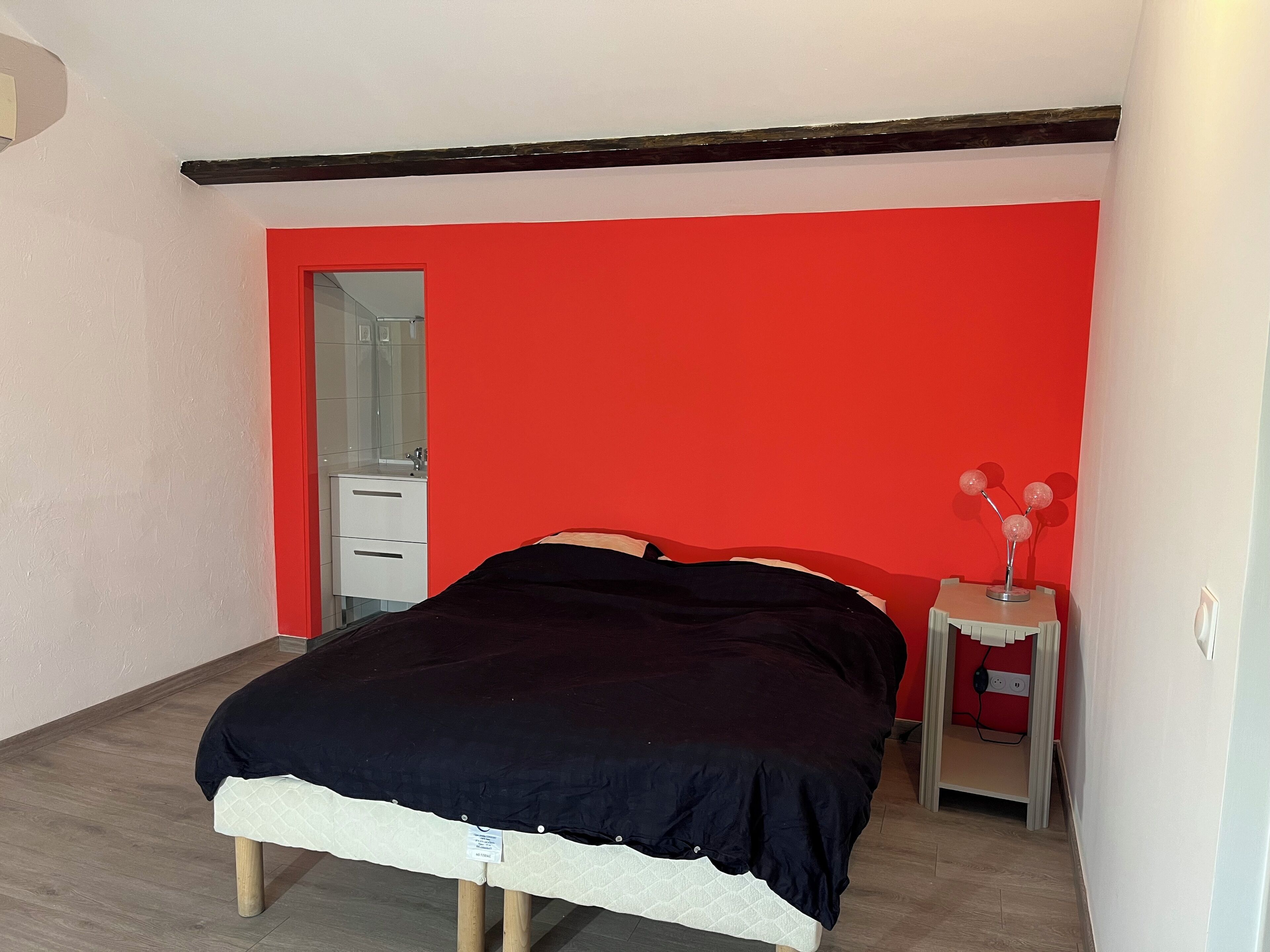 A bedroom with a bed, a small bedside table, a mirror, and a red accent wall.