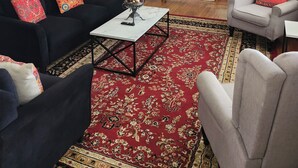 Ein Wohnzimmer mit einer schwarzen Couch, einem grauen Sessel, einem Couchtisch, einem roten Teppich mit Muster und einer Wanduhr sowie einem Deckenventilator.