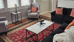 Ein Wohnzimmer mit einem roten Teppich mit Muster, einem weißen Couchtisch, einem grauen Sessel, einem schwarzen Sofa mit gemusterten Kissen und einem an der Wand befestigten Fernseher.