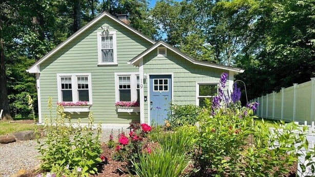 Ein zweistöckiges Haus mit einer Veranda, umgeben von einem Garten mit verschiedenen Pflanzen und Blumen.