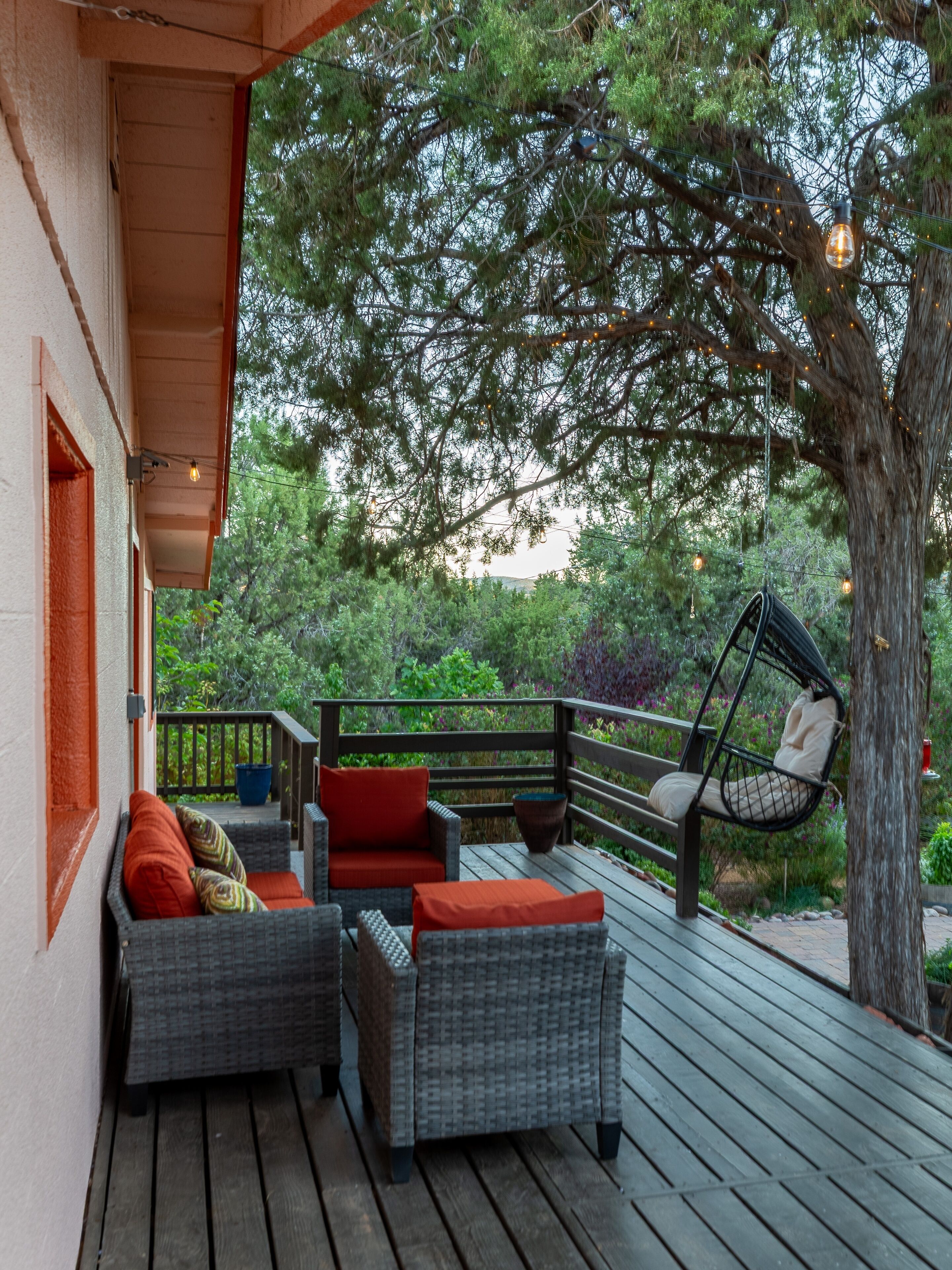 A wooden deck with wicker furniture and a hanging chair, surrounded by greenery and trees.