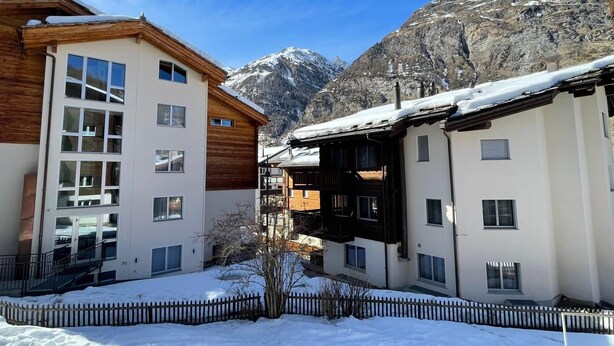 A snow-covered courtyard with modern buildings, a wooden fence, and a mountain backdrop. - Forest- finest selections (Zermatt)