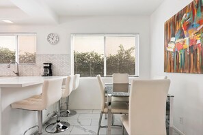 A modern kitchen with a white countertop, bar stools, a dining table with chairs, a coffee machine, and a large window with blinds. - 5bed 3bath home with piano in Los Angeles by Universal studio & Hollywood-10 (Los Angeles)