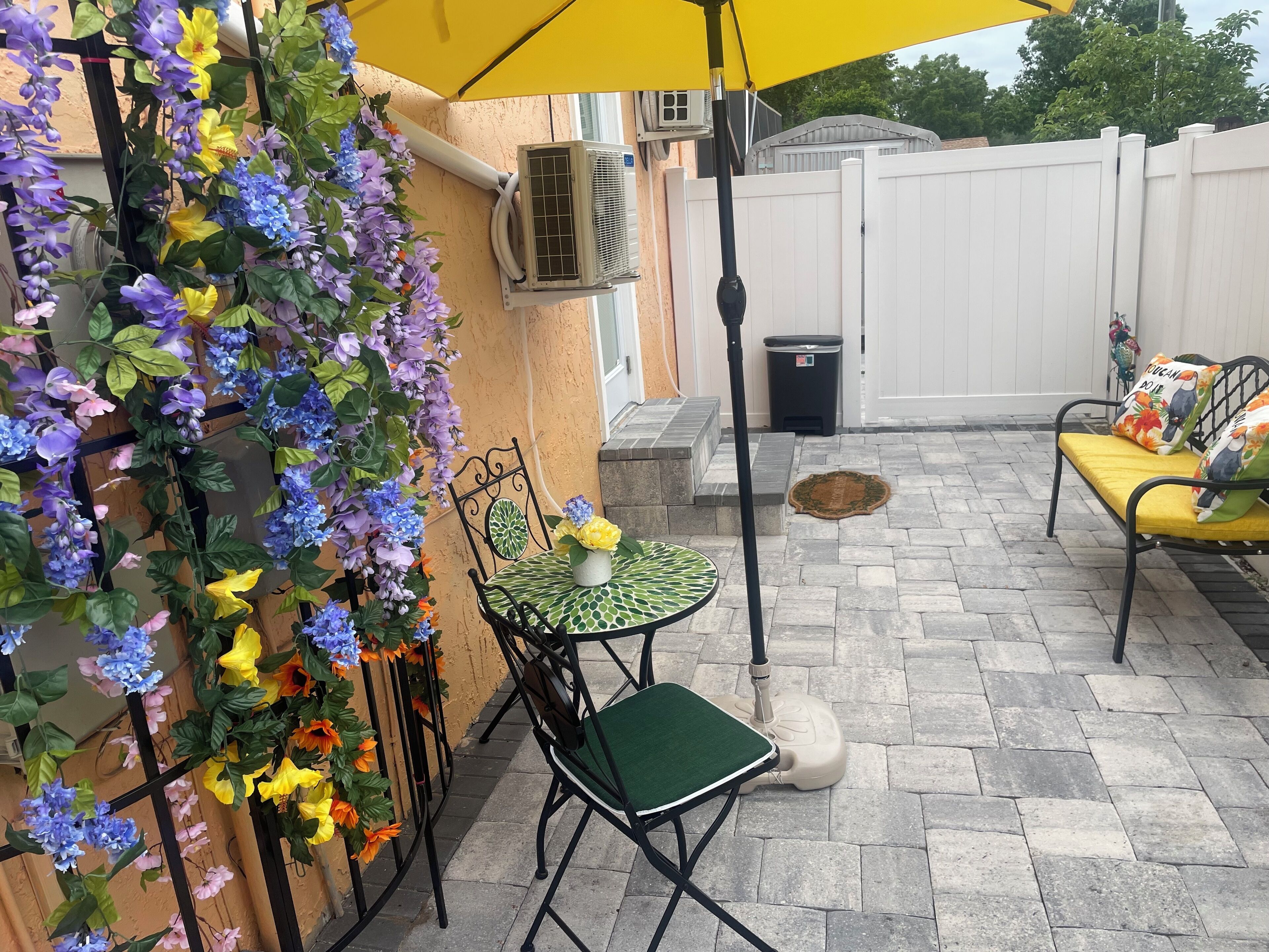 Een terras buiten met een tafel, stoelen, een gele parasol en decoratieve bloemen aan de muur.