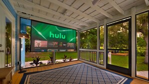 A covered outdoor area with a large screen displaying the Hulu logo, a patterned rug, and a view of a garden. - Riverside Cottage just a short walk to downtown! (Bonita Springs)