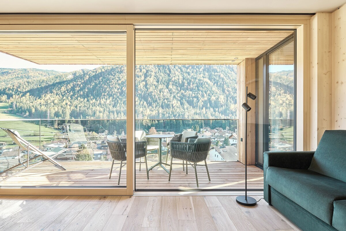 Une chambre moderne dotée d’une grande porte-fenêtre coulissante qui donne sur un balcon extérieur. Le balcon est doté d’un plancher en bois et aménagé avec une table, des chaises et un canapé.