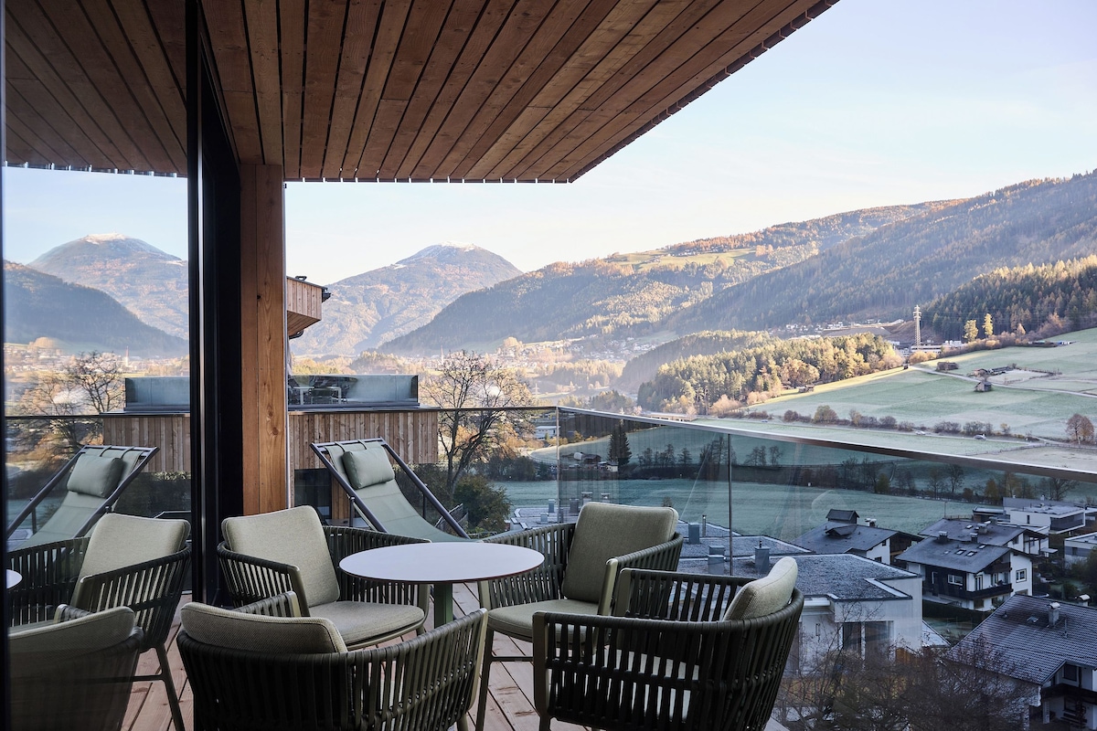 A balcony with wicker chairs and a table, overlooking a valley with mountains and a river.
