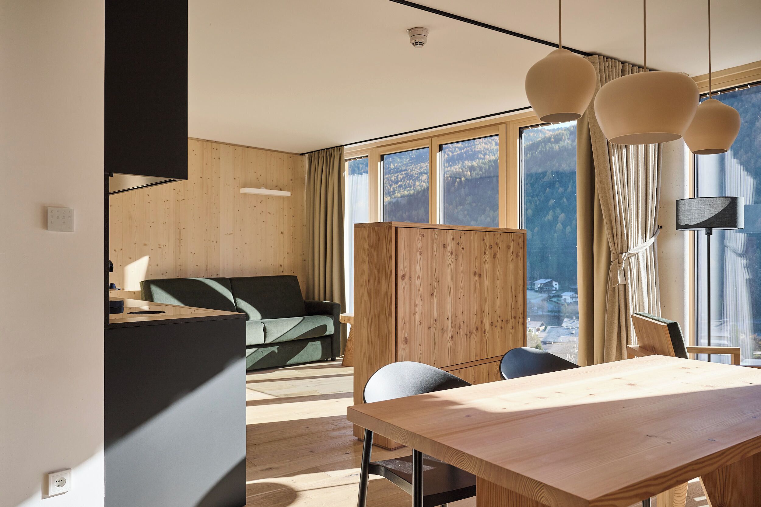 A modern interior with a wooden dining table, a sofa, and a view of mountains through large windows.