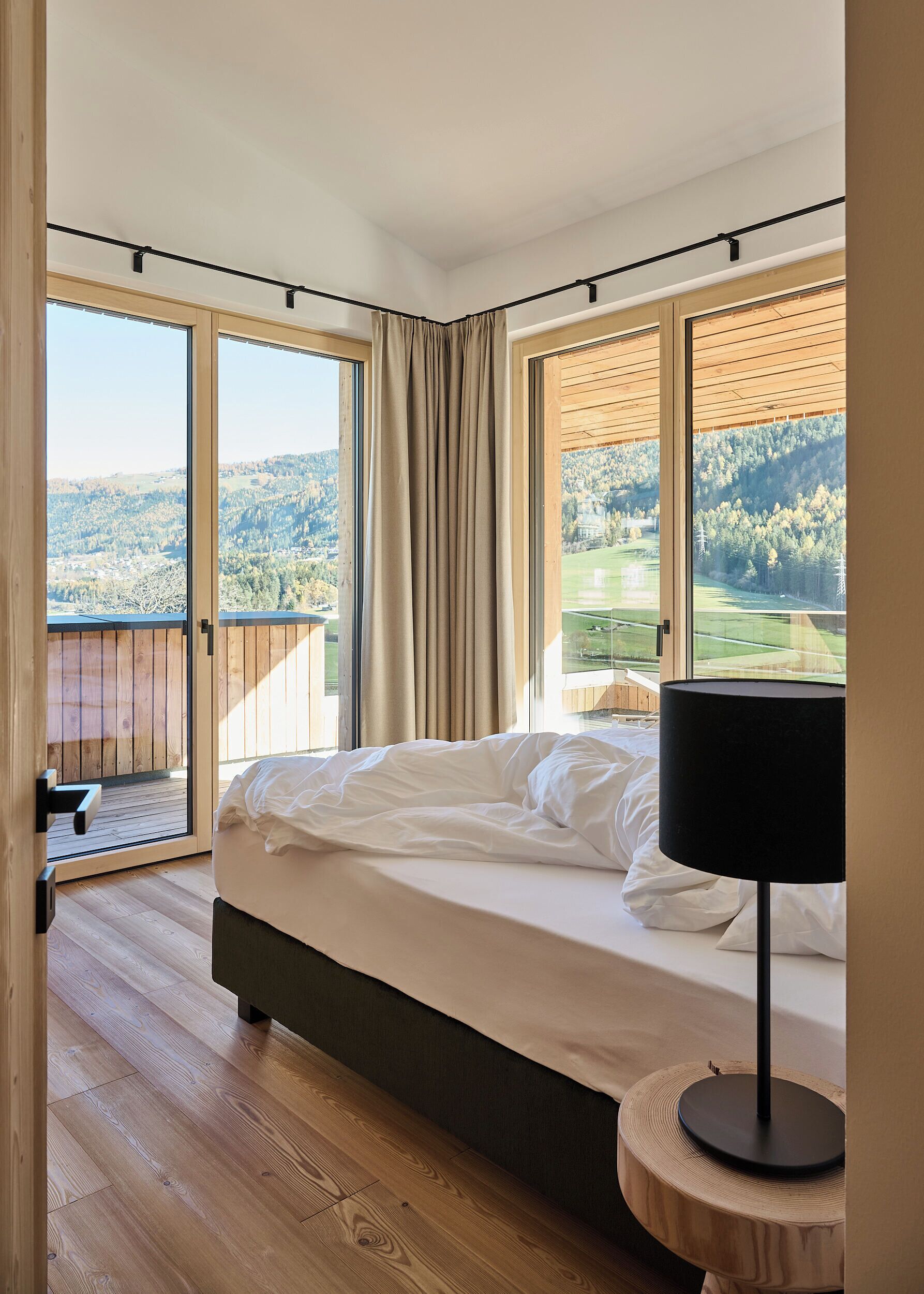Een slaapkamer met een bed, een bedlampje en uitzicht op een bebost landschap door grote ramen.