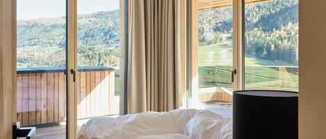Een slaapkamer met een bed, een bedlampje en uitzicht op een bebost landschap door grote ramen.