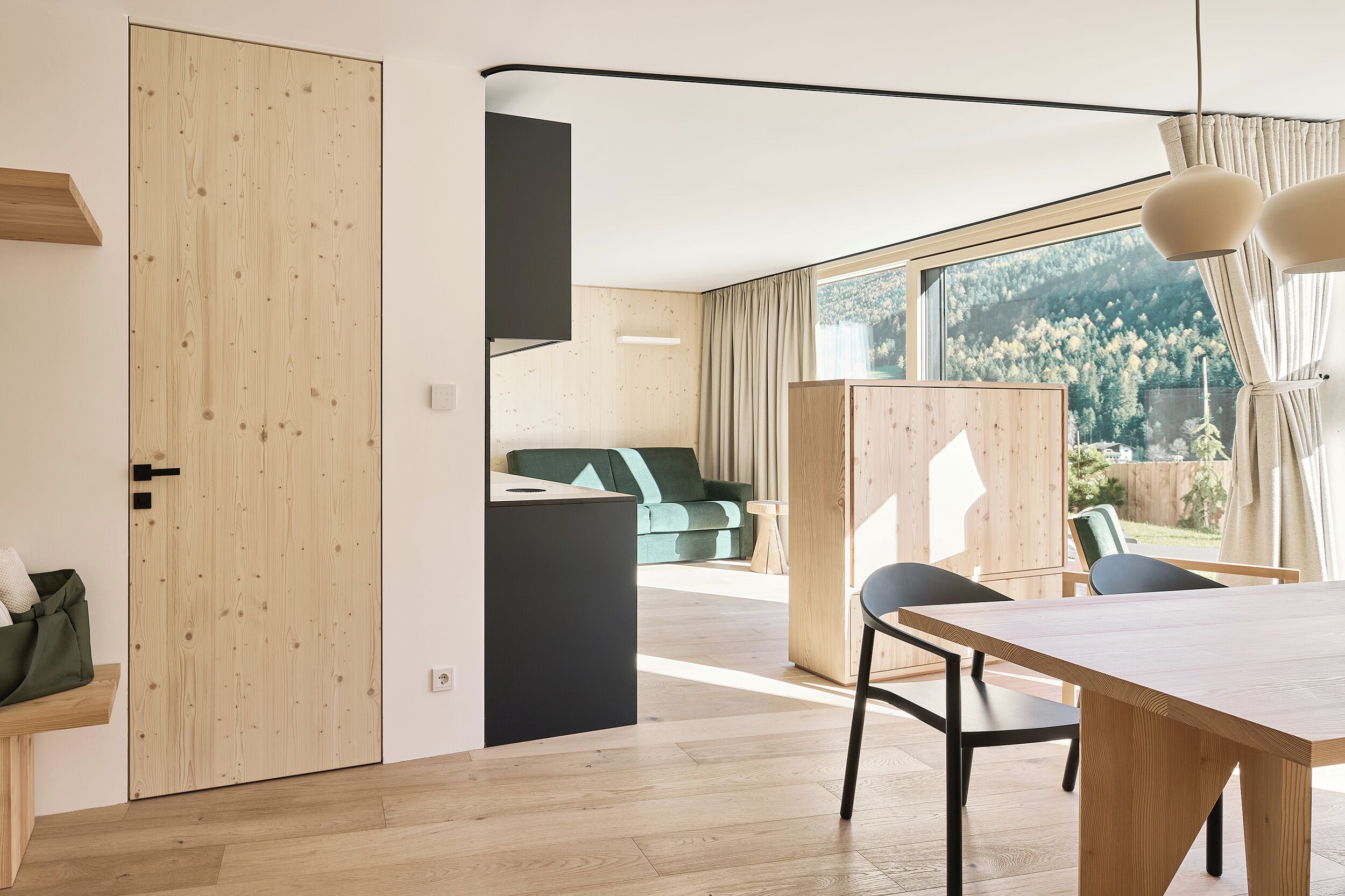 Un interno moderno con una porta in legno, una zona pranzo con tavolo e sedie, e sullo sfondo si intravedono un divano verde e un mobile in legno.
