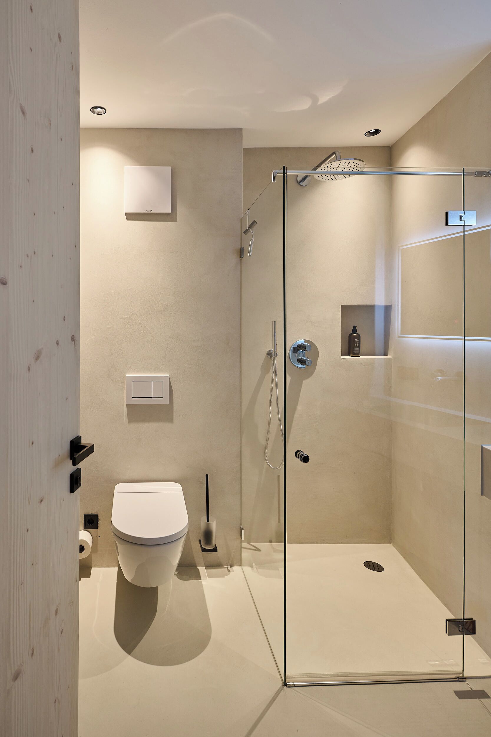 Un baño moderno con cabina de ducha de vidrio, inodoro y estante empotrado con una botella.