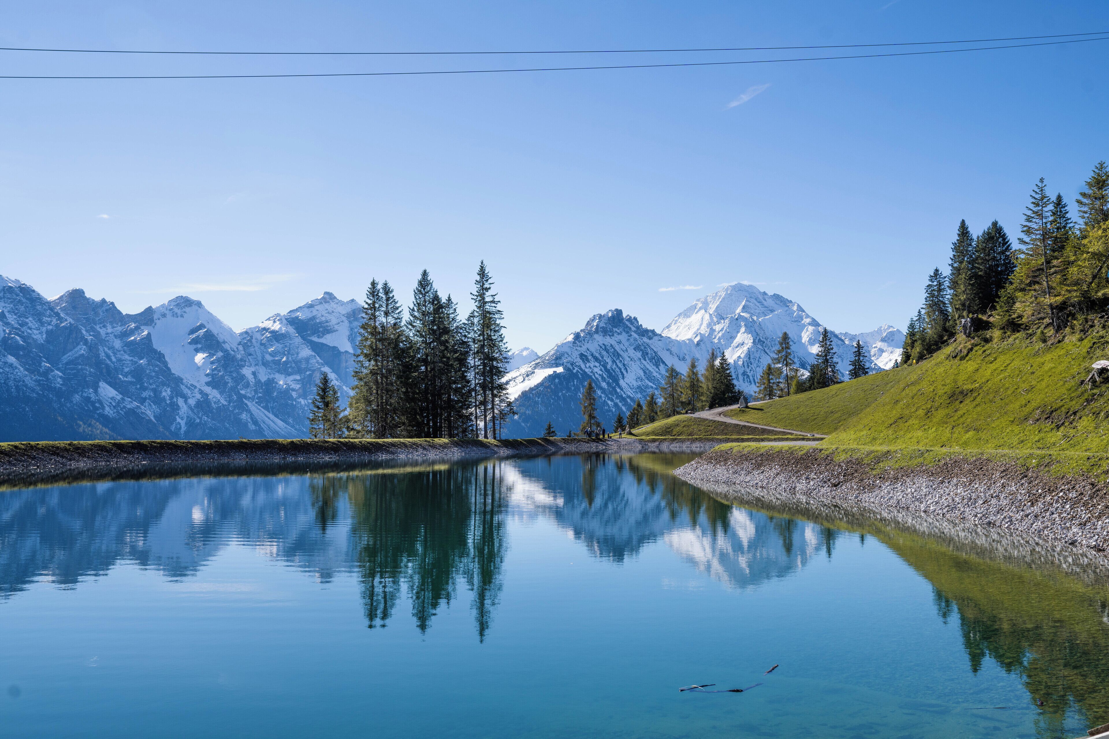 A serene mountain lake with reflections of trees and mountains, surrounded by a grassy hillside.