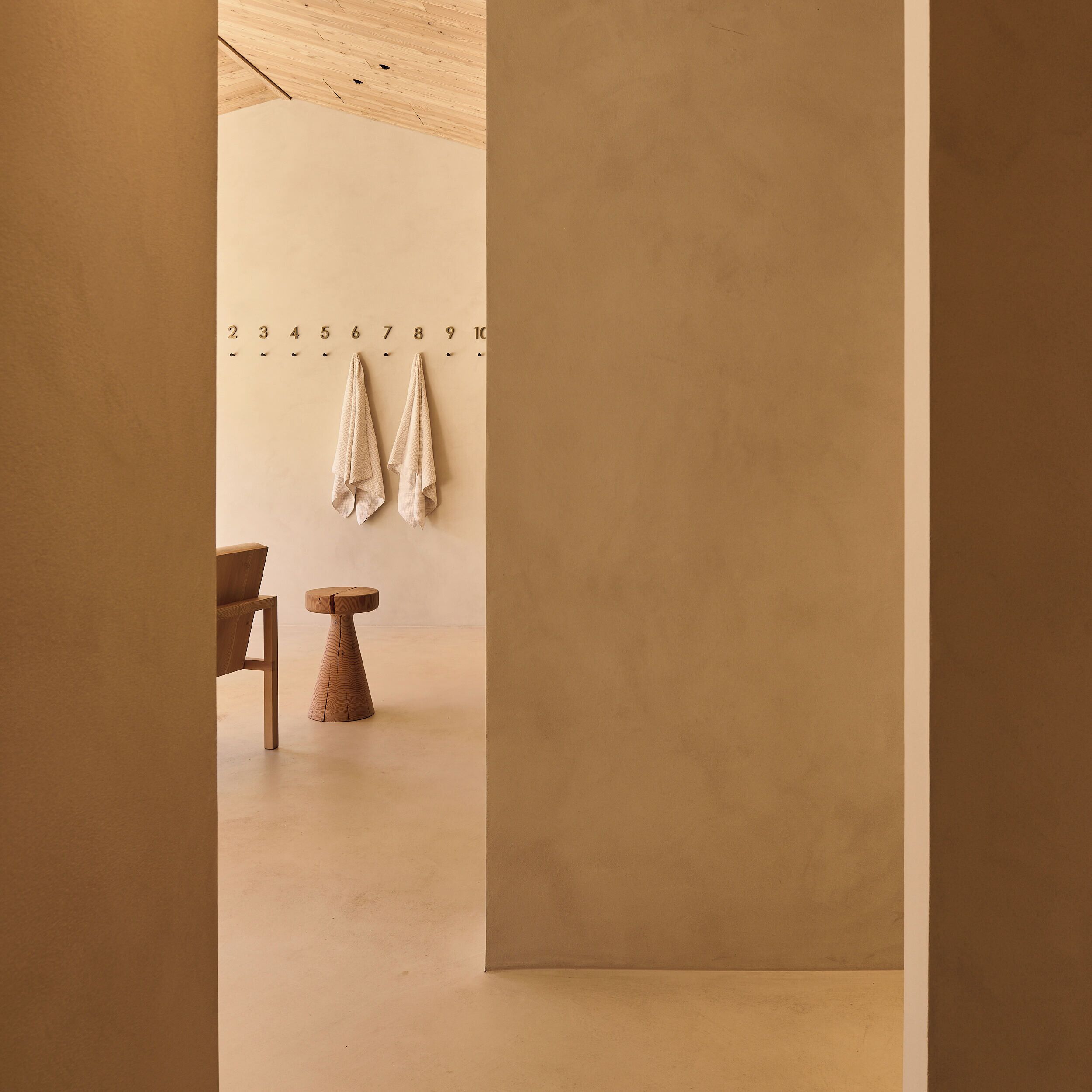 Habitación minimalista con suelo de madera, una pequeña mesa de madera y una silla. Dos toallas cuelgan de ganchos en la pared.