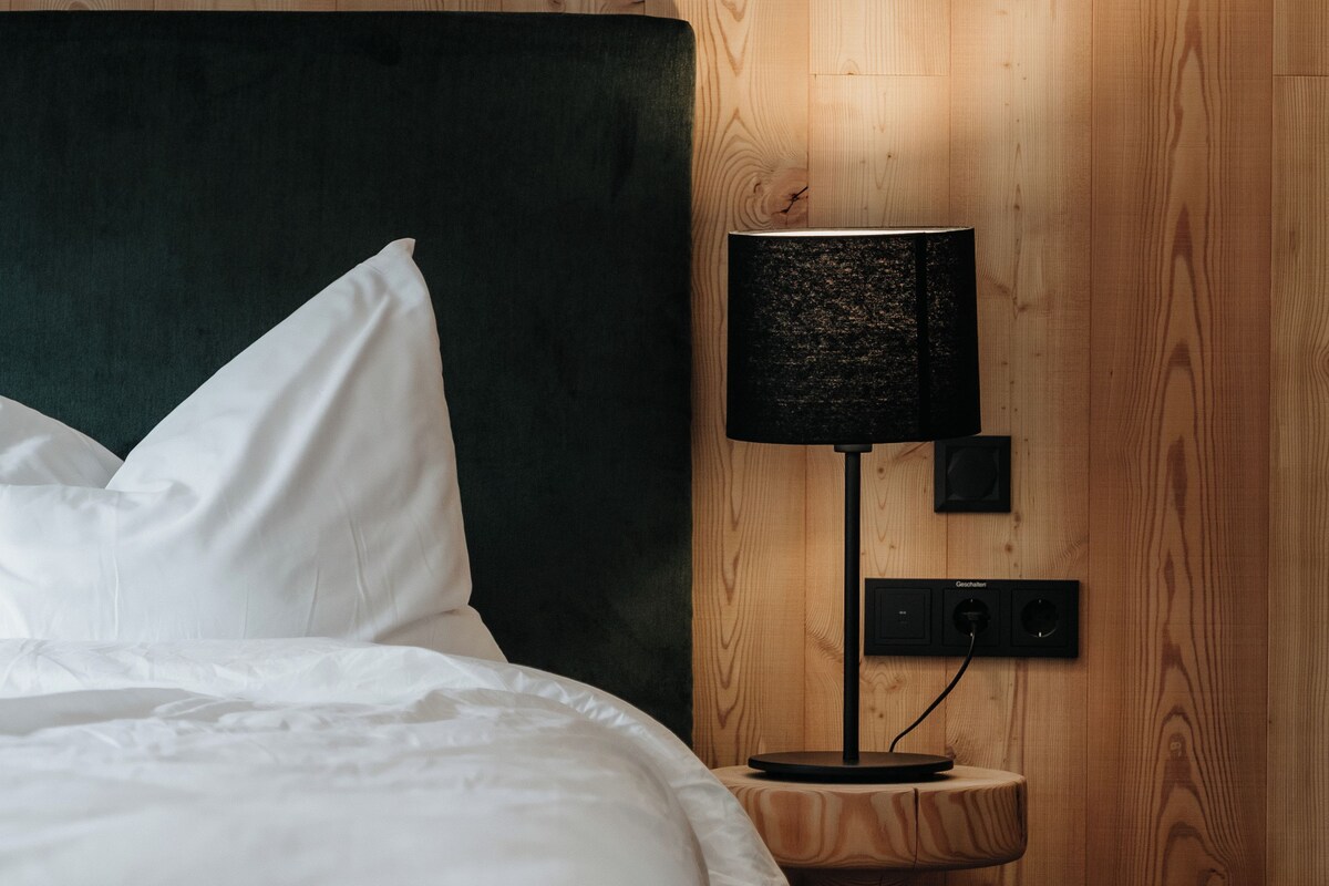 Un lit bien fait, avec du linge de lit blanc, une tête de lit sombre et une petite table de chevet en bois sur laquelle se trouve une lampe noire.