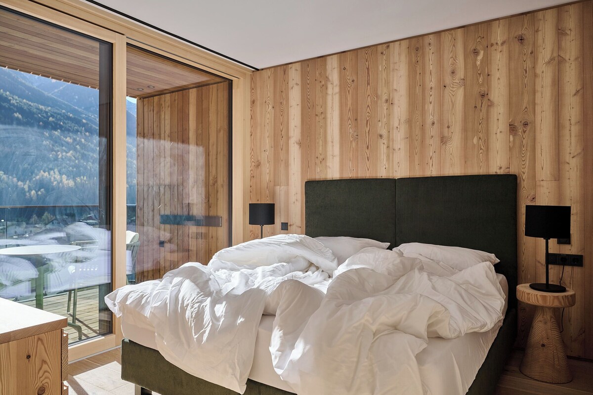 A bedroom with a large bed, wooden walls, and a view of a mountainous landscape through a glass door.