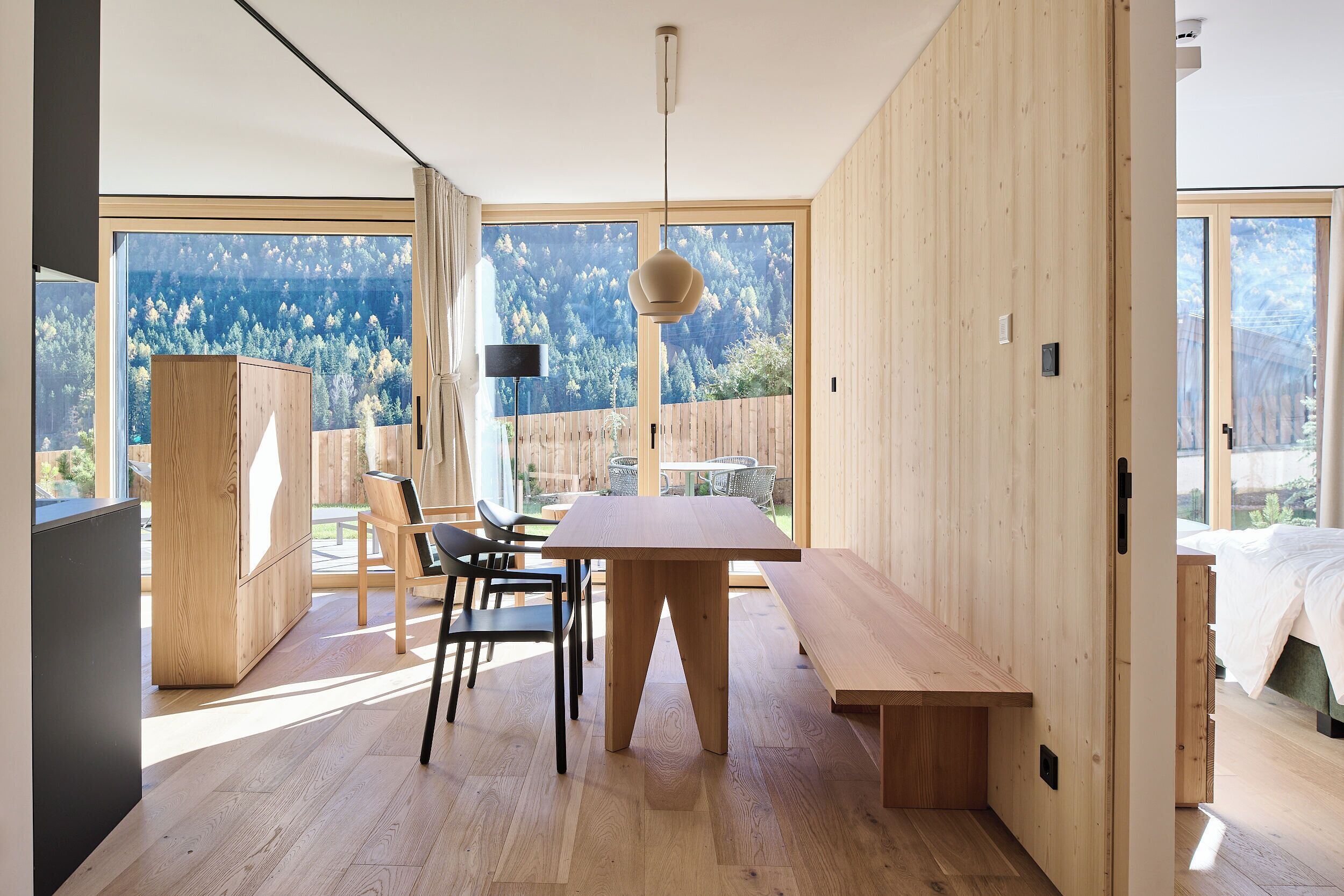 A modern interior with a wooden dining table, chairs, and a bench. There is a large window with a view of a forested area.