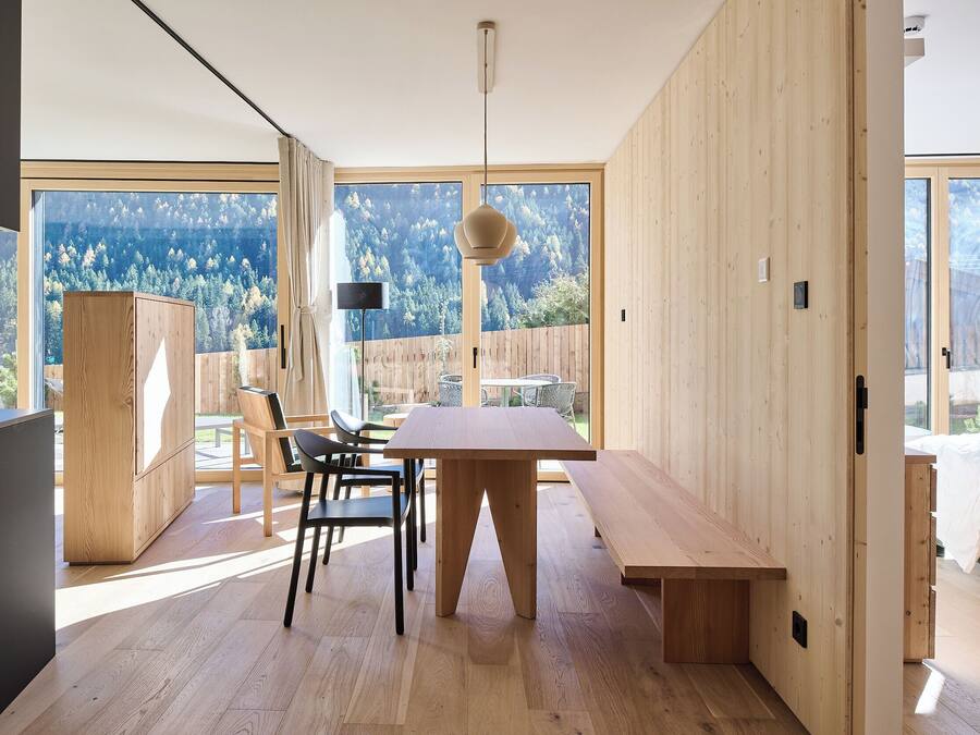 A modern interior with a wooden dining table, chairs, and a bench. There is a large window with a view of a forested area.