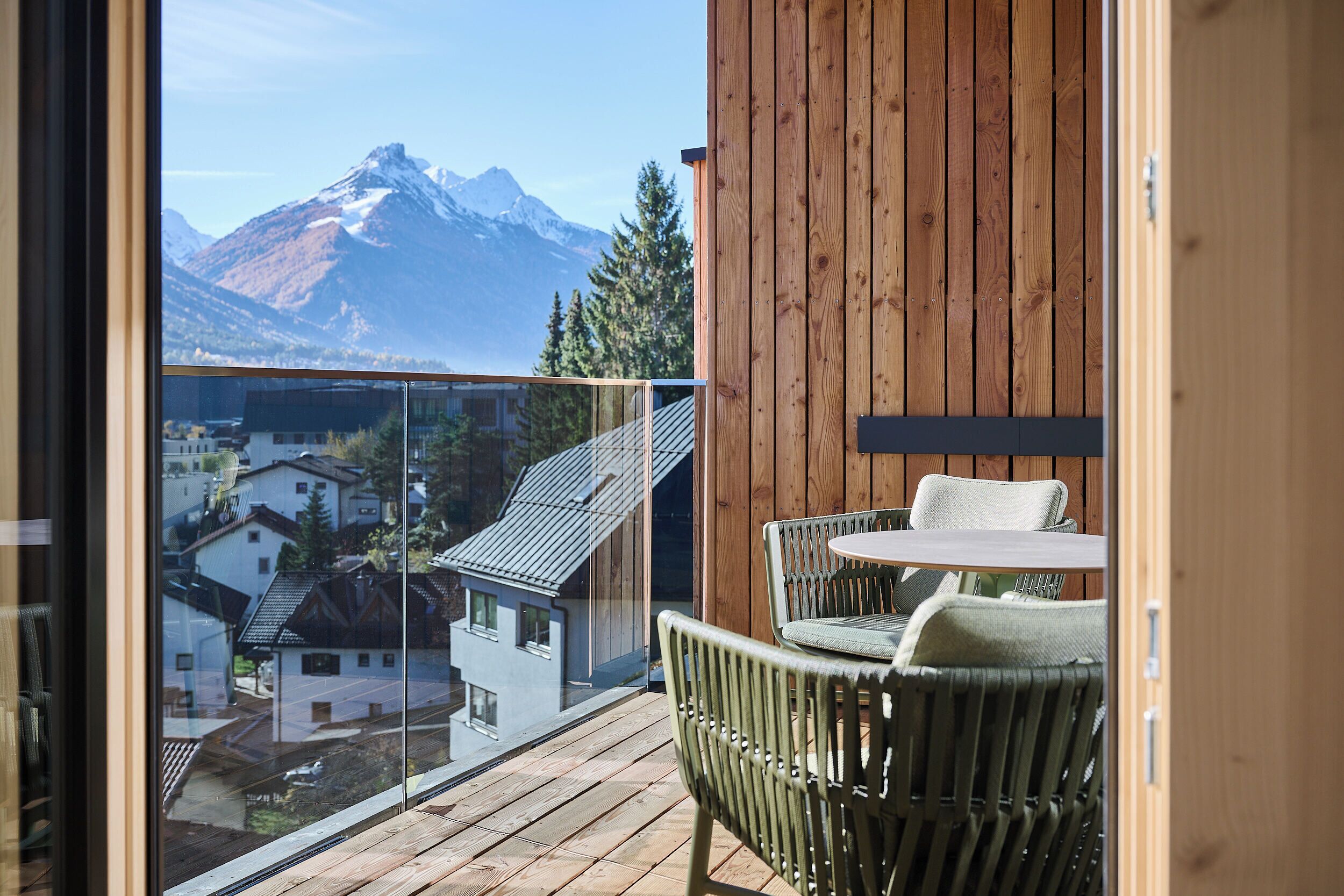 Un balcone con pavimento in legno e sedie in vimini, con vista su una montagna e edifici residenziali.