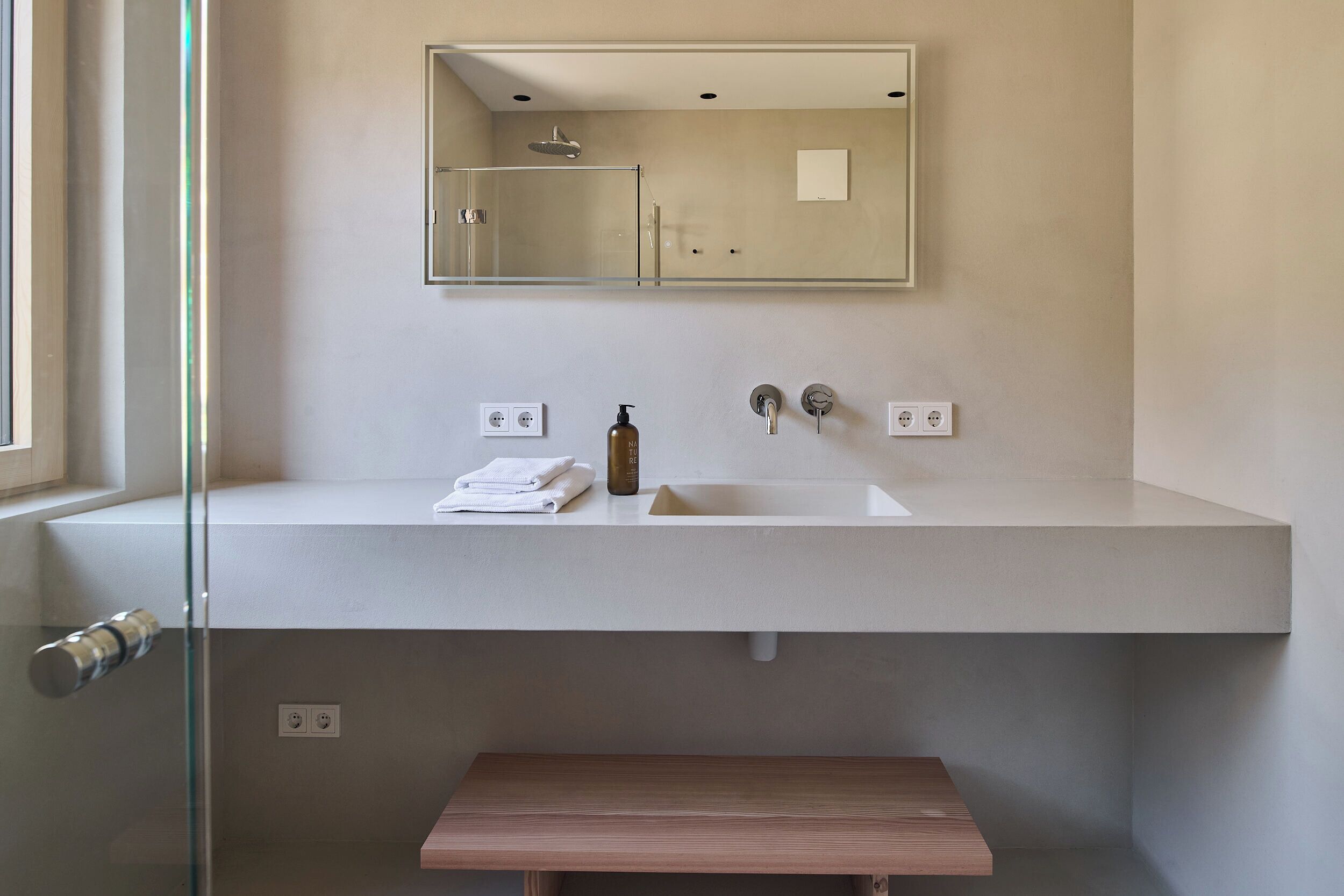Ein modernes Badezimmer mit einem weißen Waschbecken, einer Duschkabine aus Glas und einer hölzernen Sitzbank.