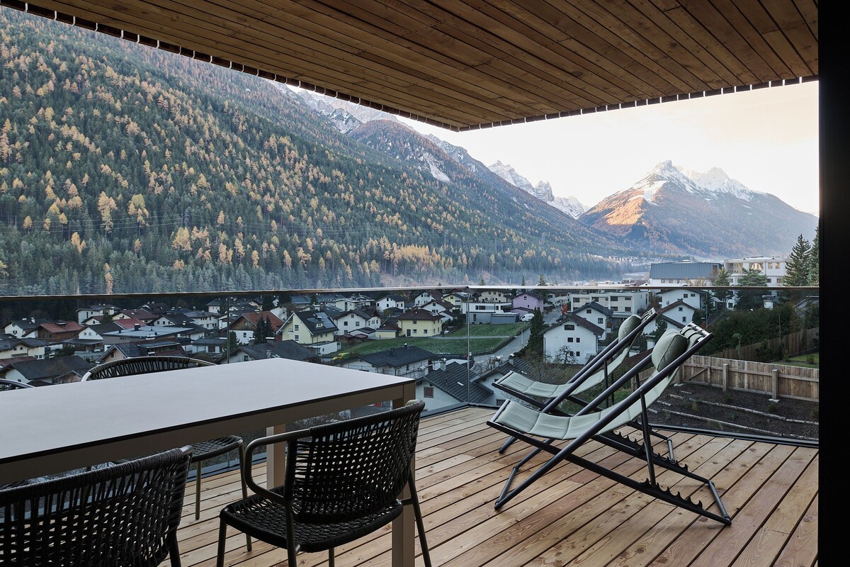 Une terrasse en bois avec une table et des chaises, offrant une vue sur un paysage montagneux avec des maisons situées dans la vallée en contrebas.