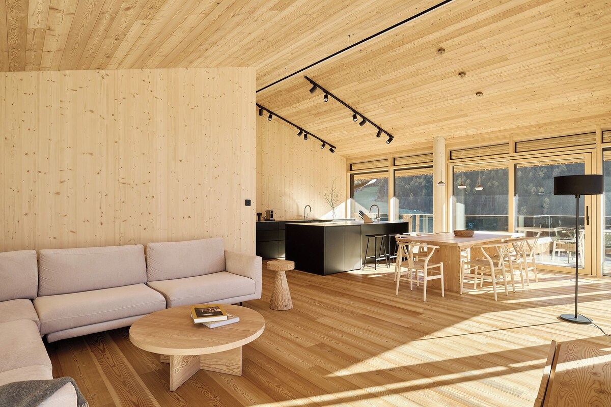 A modern interior with a wooden ceiling, a sofa, a round coffee table, a dining area, and a kitchen with large windows.