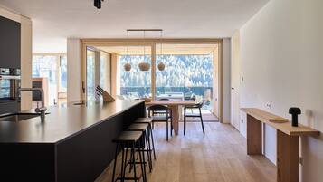 A modern kitchen with a large island, bar stools, a dining table, and large windows offering a view of trees.