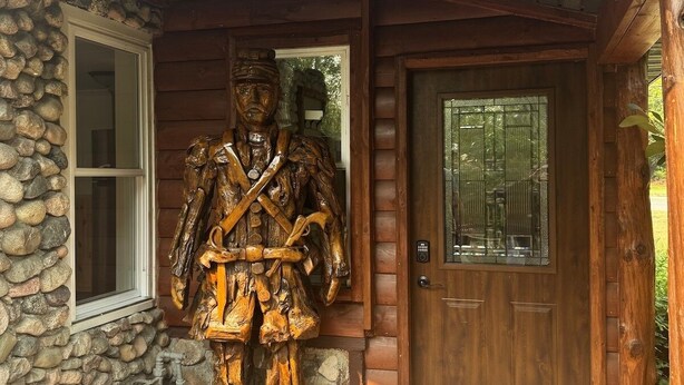 Une cabane rustique avec une statue en bois représentant une figure sur le porche, un mur en pierre et une porte en bois dotée d’une fenêtre.