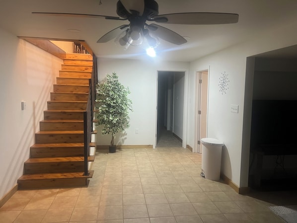 Una escalera de madera con peldaños que conduce a una plataforma, un ventilador de techo, una planta en maceta y un pasillo con una puerta al final.