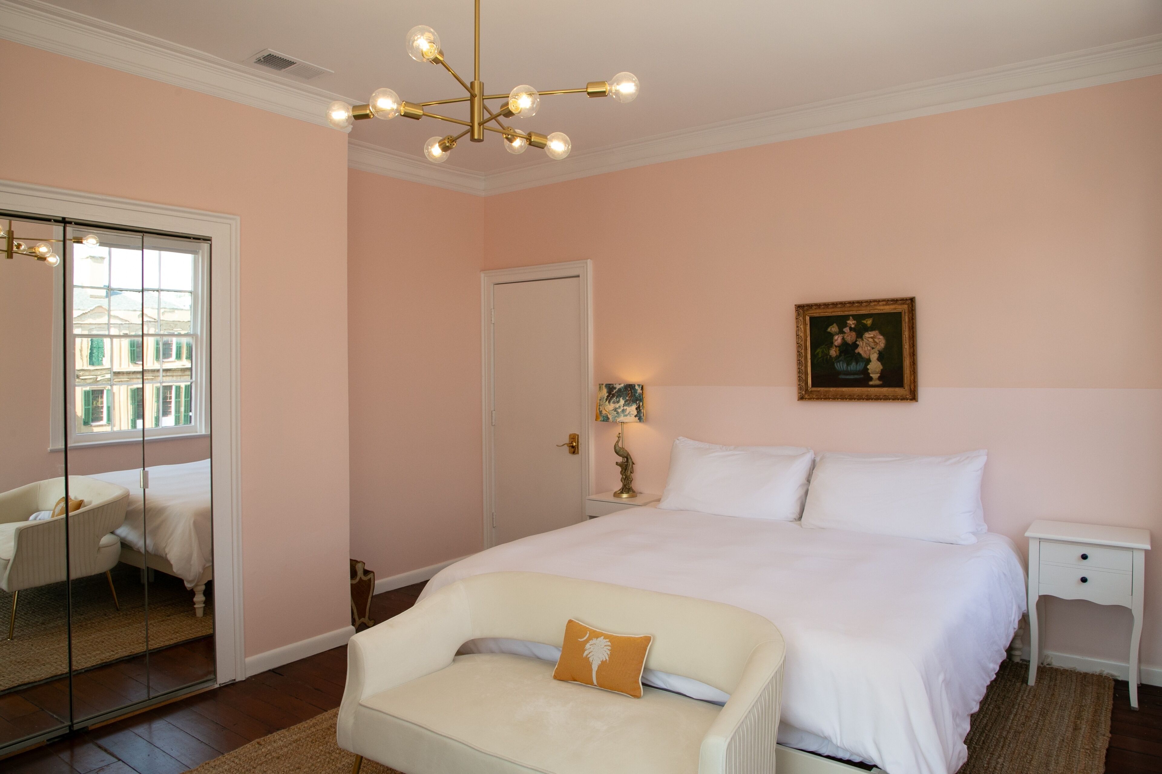 Un dormitorio con una cama grande, una mesita de noche blanca, una mesilla auxiliar, una silla, un espejo y una puerta.