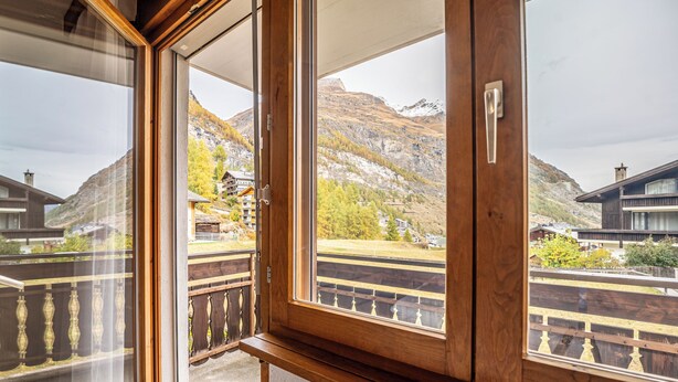 A wooden-framed window with a view of a mountainous landscape and a building with a balcony. - Fabi s Horu Lodge (Zermatt)