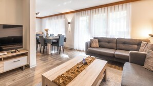 A modern living room with a flat-screen TV, a grey sofa, a wooden coffee table, and a dining area with a table and chairs. - Fabi s Horu Lodge (Zermatt)