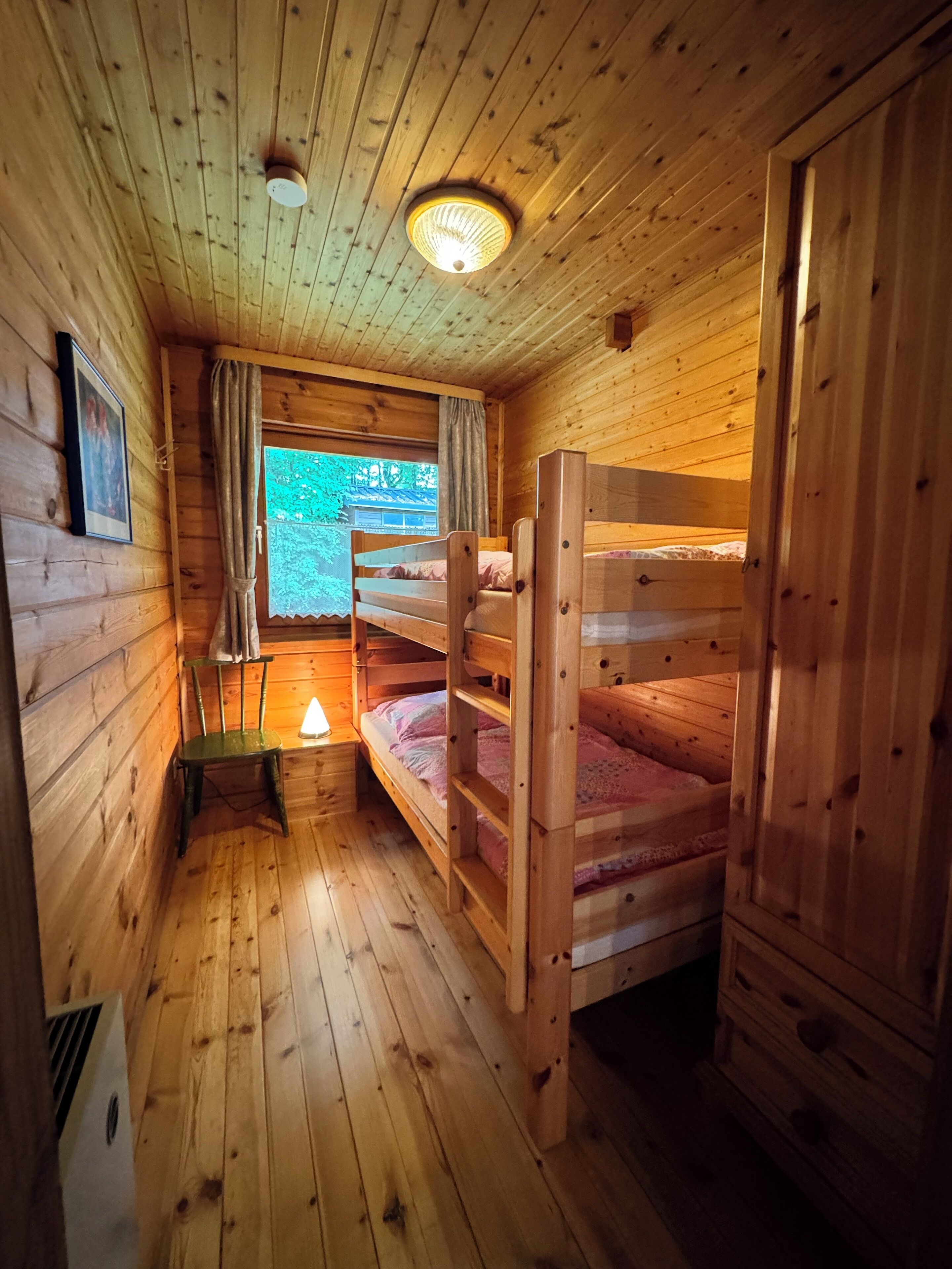 A wooden cabin room with bunk beds, a window with curtains, a framed picture on the wall, and a small table with a lamp.