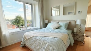 A bedroom with a large window, a bed with a quilt, two bedside tables with lamps, and a wall-mounted thermostat. - Roger & May's Cabin in Vancouver (Vancouver)