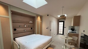 Une chambre moderne avec une lucarne, un lit, une petite table ronde et, au fond, un coin cuisine.