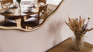 Et spiseområde med et træbord dækket til fire personer, en vase med tørrede blomster og et guldrammet spejl der reflekterer rummet.