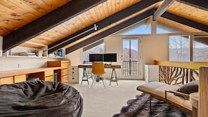 Un dormitorio acogedor en el ático, con techo de madera, un escritorio con computadora, un sofá y un amplio ventanal con vistas al exterior.