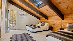 Un dormitorio acogedor con techo inclinado, cama con edredón blanco y almohadones oscuros, alfombra mullida y un cuadro en la pared.