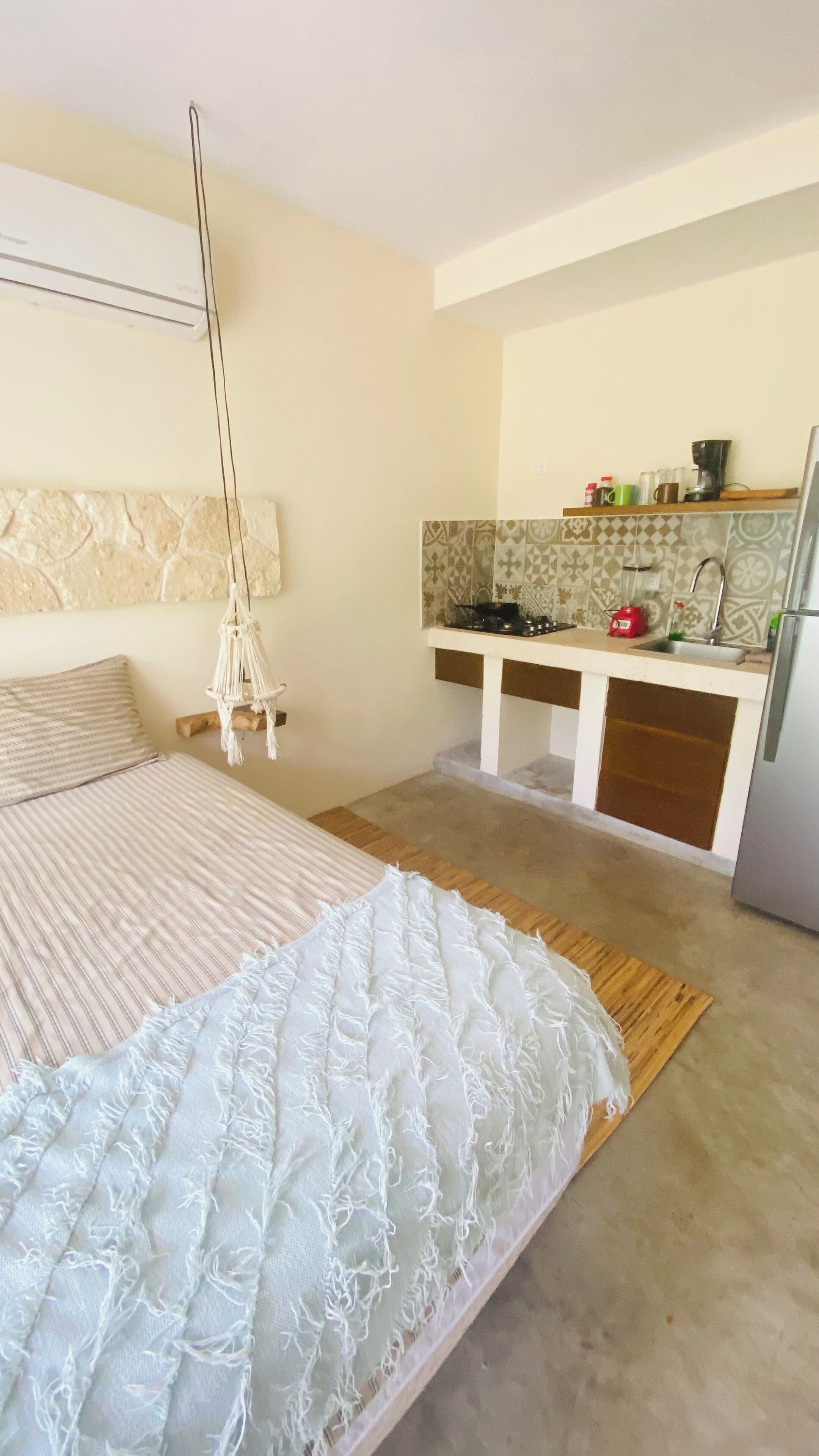 A bedroom with a bed, a hanging macrame light, a kitchenette with a stove, and a refrigerator.