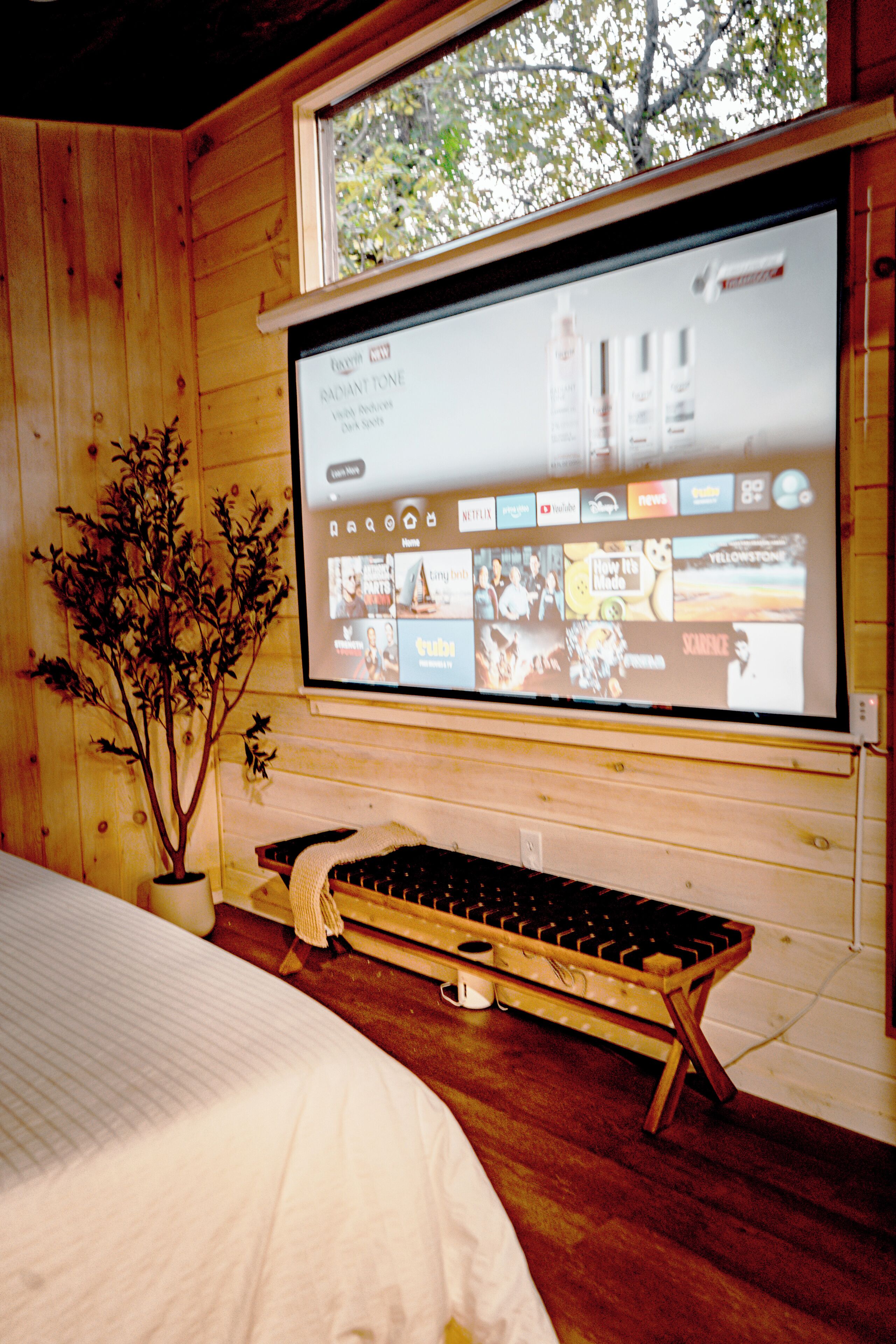 Ein Zimmer in einer Holzhütte mit einem Bett, einer Projektionsleinwand, die einen Film zeigt, und einer kleinen Pflanze.