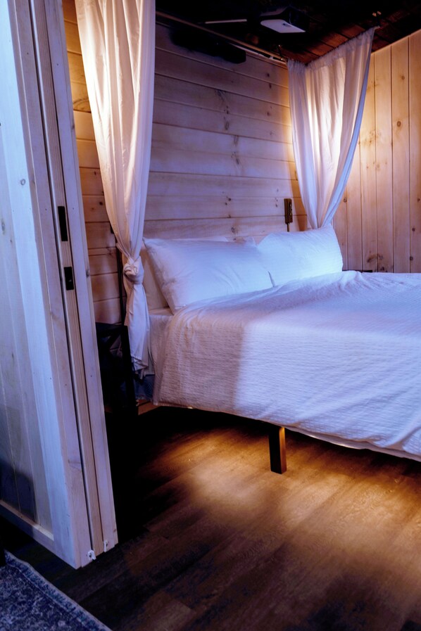 A wooden cabin bedroom with a bed, white curtains, and a wooden floor.