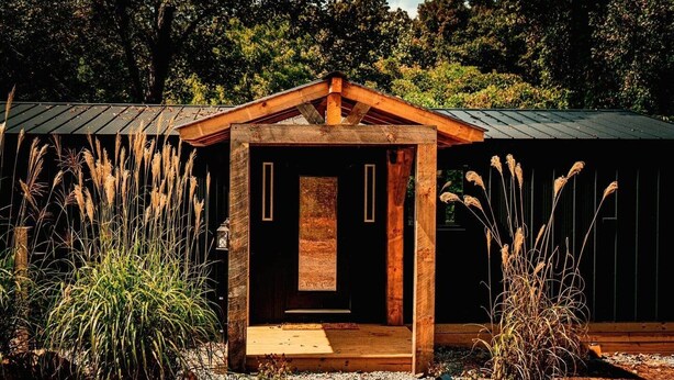 A rustic wooden cabin with a central door and a small porch area, surrounded by tall grasses and trees. - The Laurel  A private retreat for two! Scenic mornings. (Fannin County)