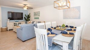 Una sala de estar moderna con un sofá, una mesa de comedor y sillas. Hay un televisor, un ventilador de techo y una lámpara decorativa.
